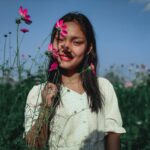 Positive young ethnic female in white dress holding stems of flowers putting buds to face while standing among blooming plantation against blue sky in sunny day
